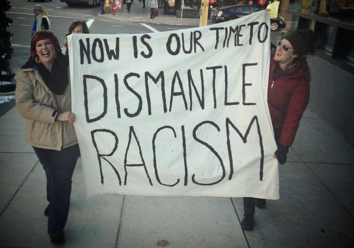 dismantleracism two people walking and holding a large sign that reads: now is our time to dismantle racism