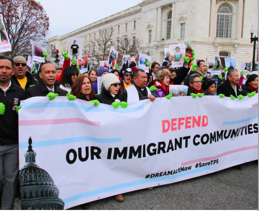 People are marching holding a large banner that reads "defend our immigrant communities"